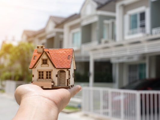 A hand holding a model house in front of a suburb - How Long Does It Take To Close On A House? - DG Pinnacle Home Loans - Miami Mortgage Funding Lending Company