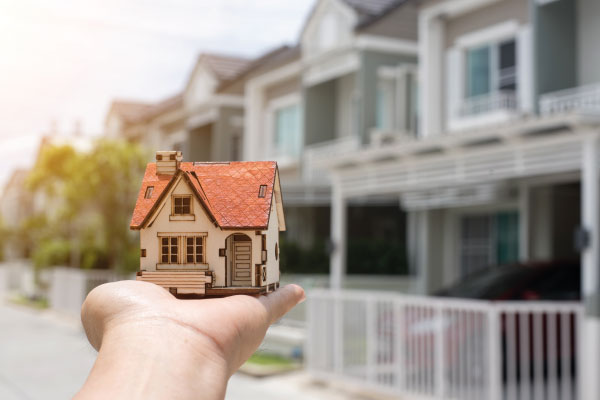 A hand holding a model house in front of a suburb - How Long Does It Take To Close On A House? - DG Pinnacle Home Loans - Miami Mortgage Funding Lending Company
